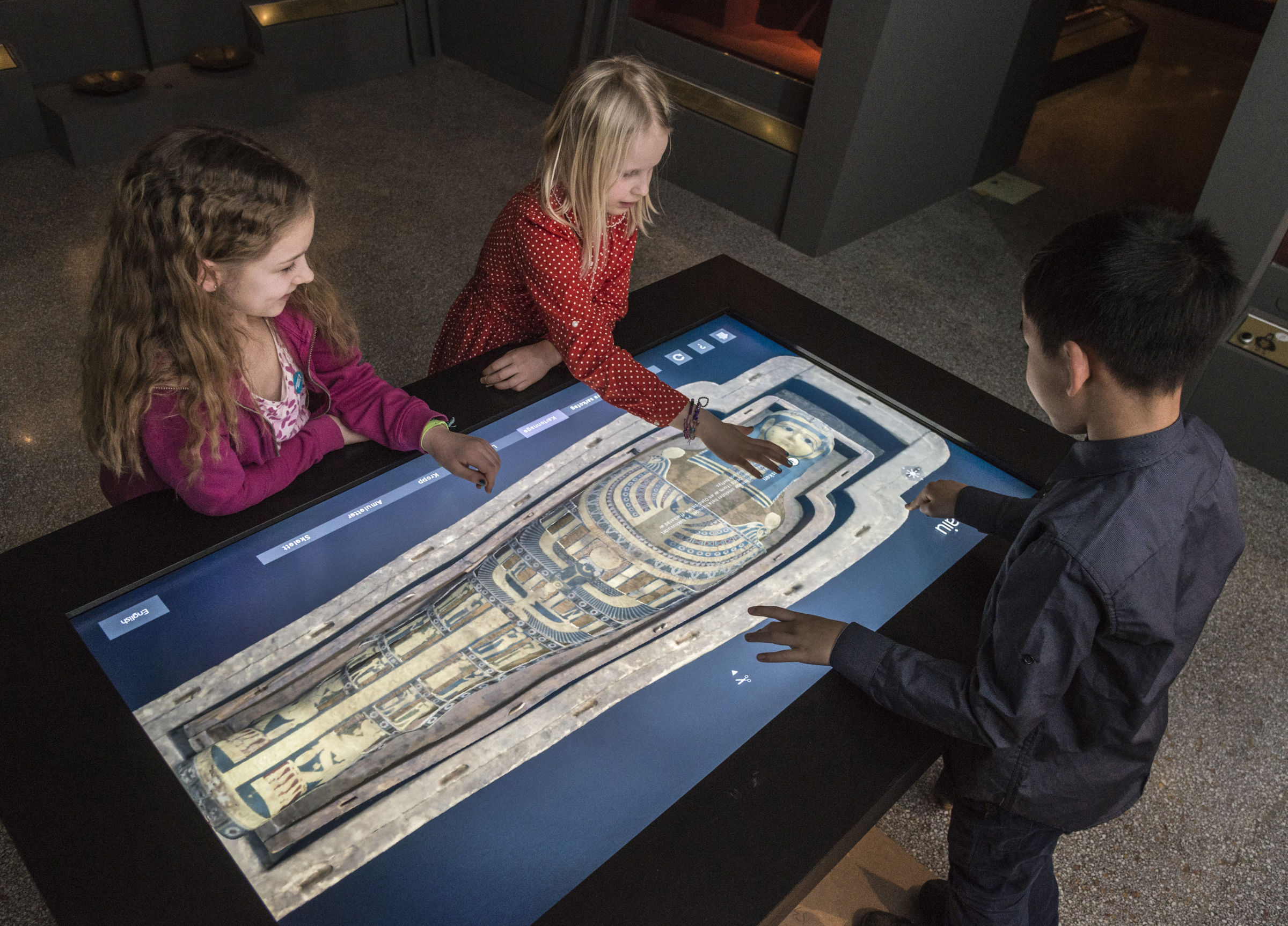 Three kids using a large touch table to look at a 3D scan of a sarcophagus